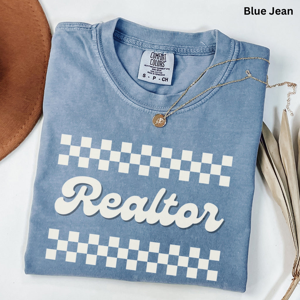 A blue t-shirt with a checkered pattern and the word "Realtor" printed on it, along with a necklace and some decorative leaves.