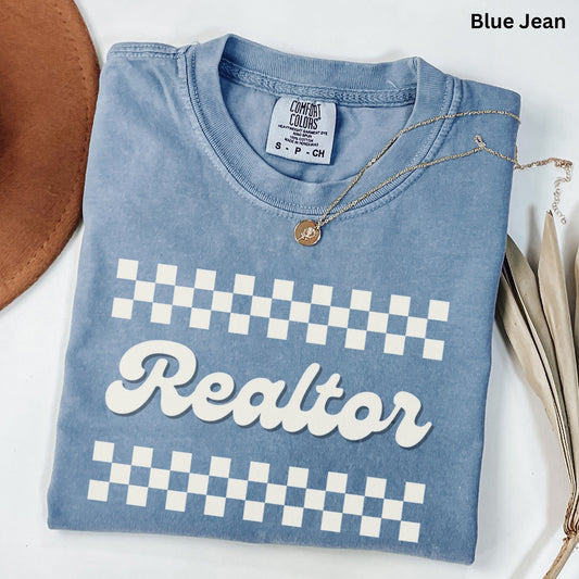 A blue t-shirt with a checkered pattern and the word "Realtor" printed on it, along with a necklace and some decorative leaves.