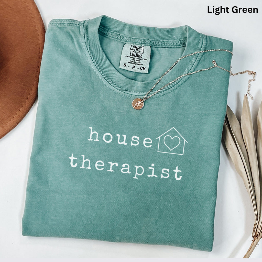 A light green t-shirt with the text "house therapist" printed on it, along with a heart symbol and a house icon. The t-shirt is folded and placed on a white surface, with a brown hat and some dried leaves nearby.