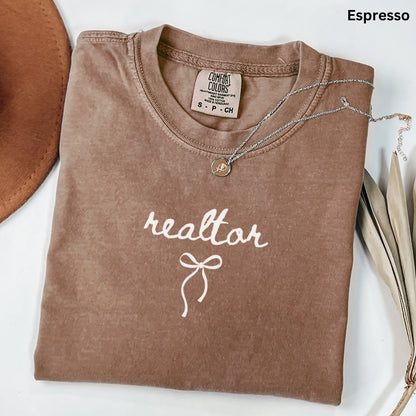 a brown t-shirt with the word "realtor" printed on it, along with a small charm or pendant. The t-shirt is folded and placed on a white surface, with a brown hat and some leaves visible in the background.
