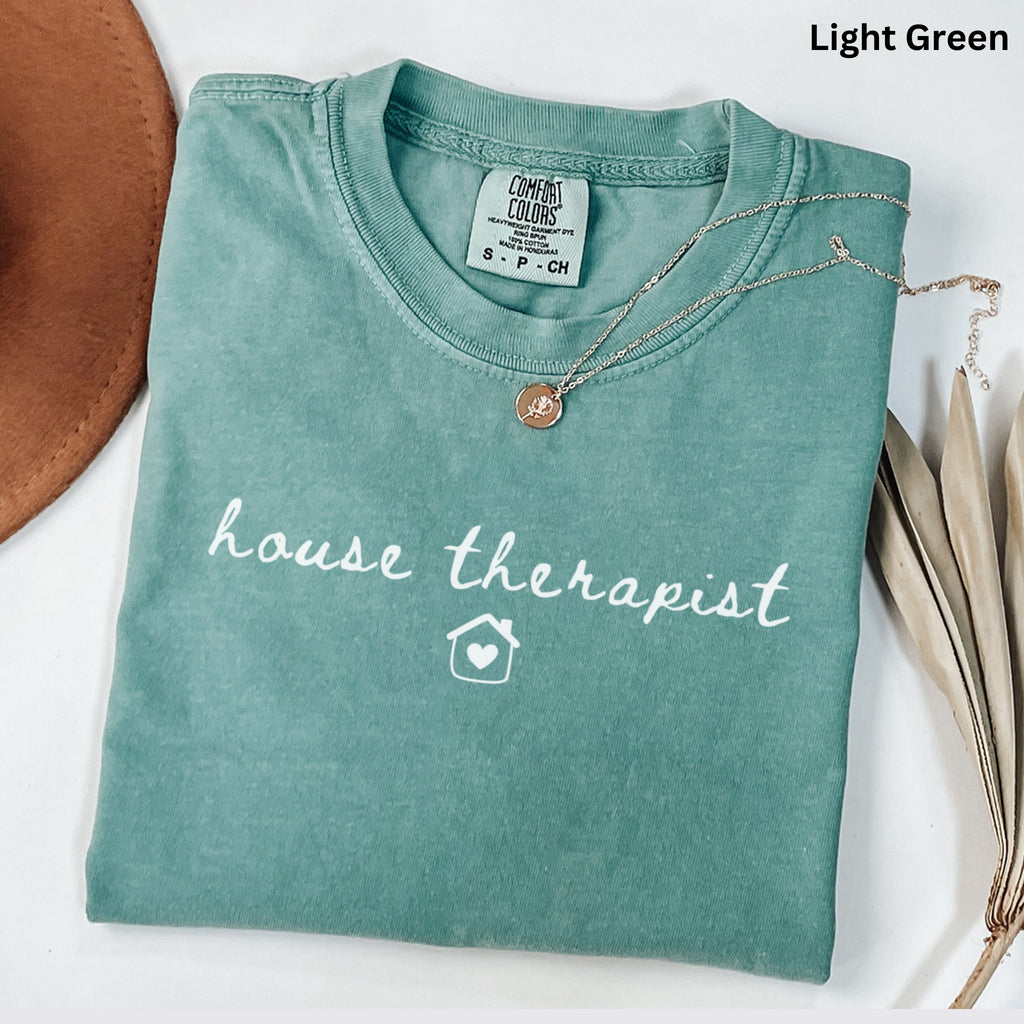 A light green t-shirt with the text "house therapist" printed on it, along with a small house icon. The t-shirt is folded and placed on a white surface, with a brown hat and some dried leaves nearby.