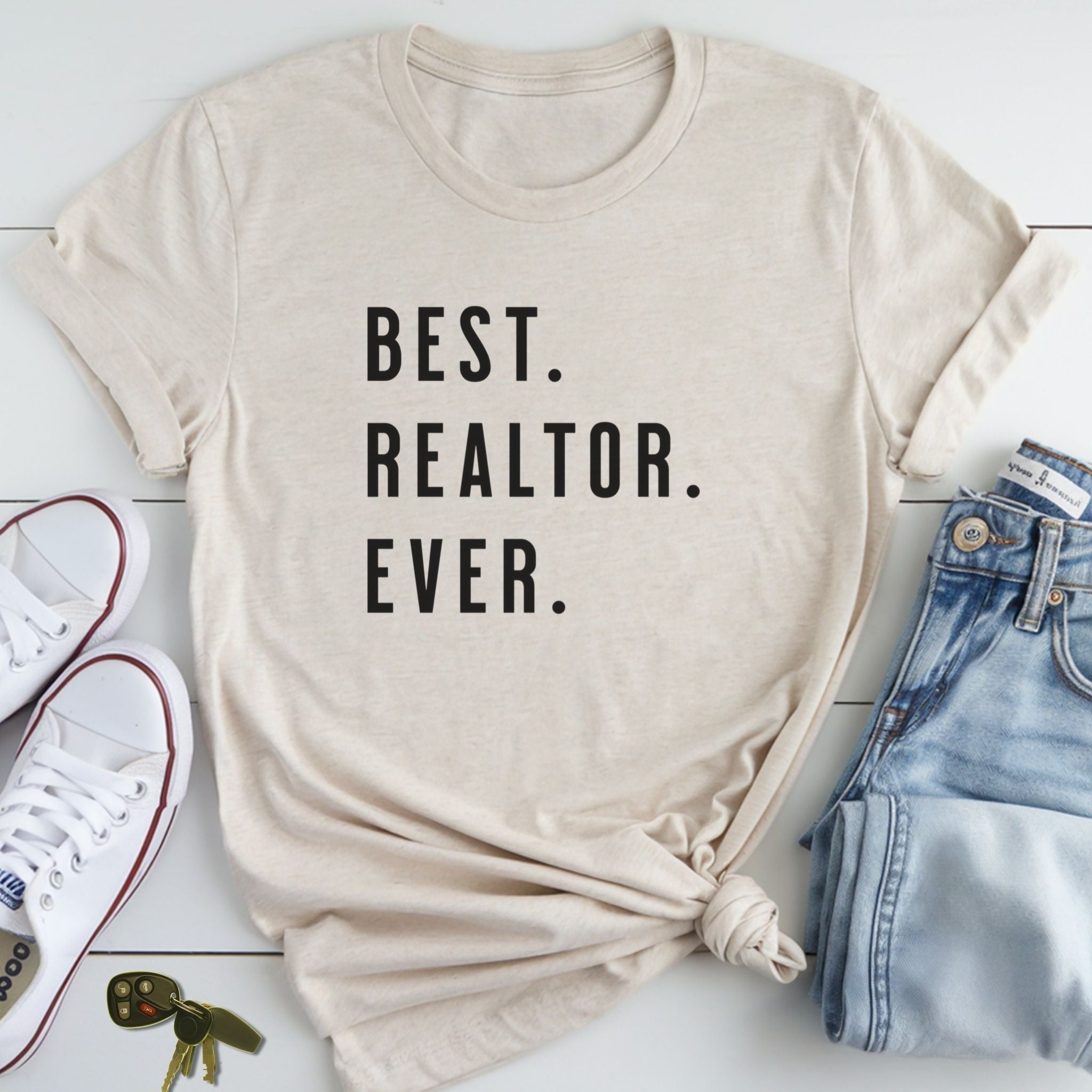 A beige t-shirt with the text "BEST REALTOR. EVER." printed on it, along with a pair of jeans and white sneakers.