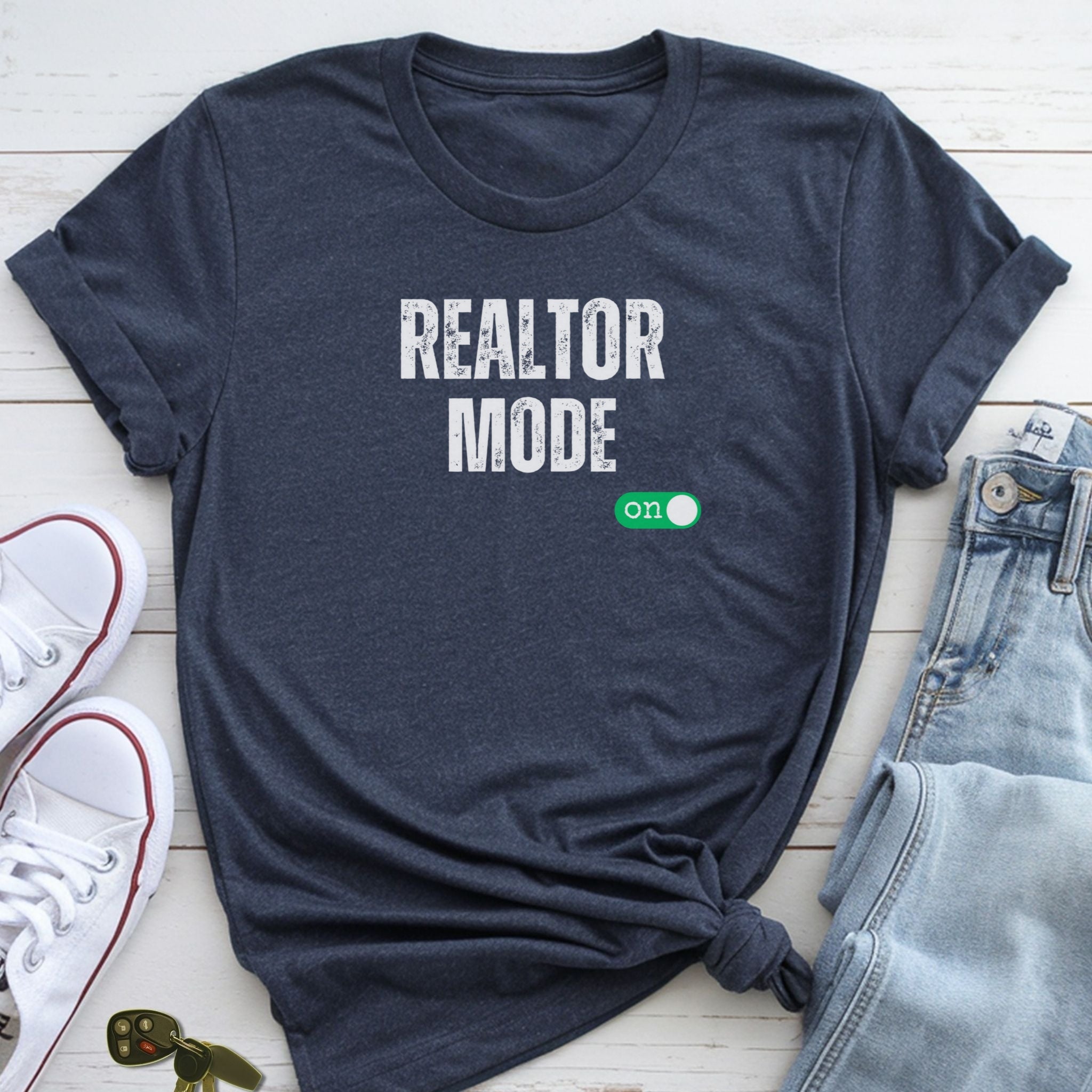 A dark blue t-shirt with the text "REALTOR MODE" printed on it, along with a green button icon, is displayed on a wooden surface alongside a pair of jeans and white sneakers.