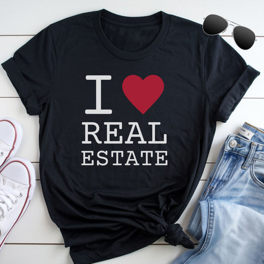 A black t-shirt with the text "I ❤️ REAL ESTATE" printed on it, along with a red heart symbol. The t-shirt is placed on a white surface, and there are a pair of sunglasses and a pair of jeans visible in the image.