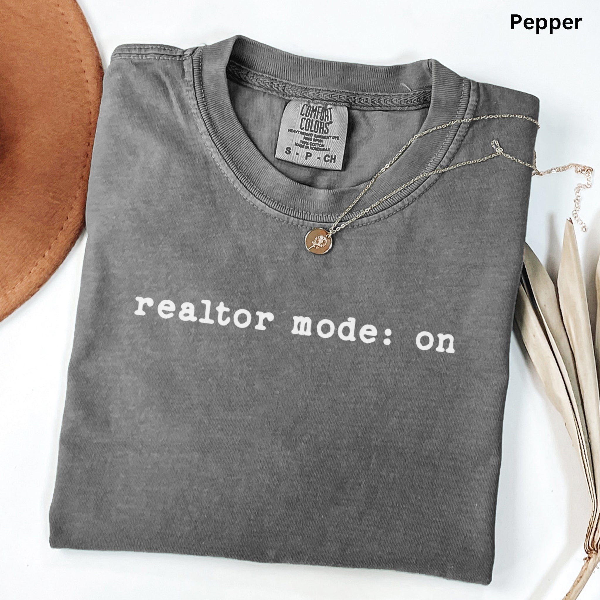 A gray t-shirt with the text "realtor mode: on" printed on it, along with a necklace and some leaves.