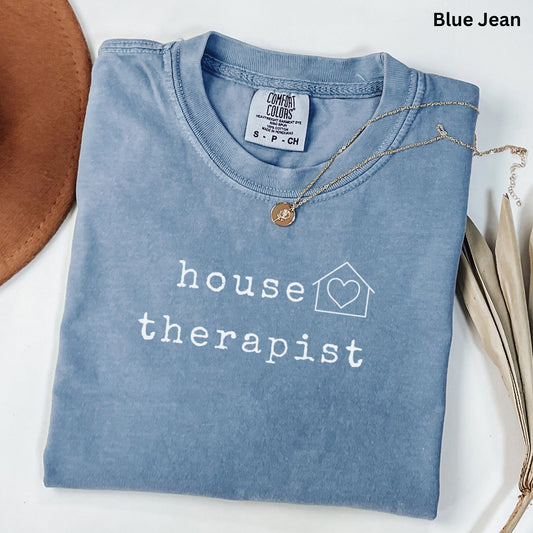 A blue t-shirt with the text "house therapist" printed on it, along with a heart symbol inside a house. The t-shirt is folded and placed on a white surface, with a brown hat and some leaves nearby.