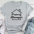 A gray t-shirt with the text "Property Manager" printed on it, along with a graphic of a house. The t-shirt is laid out on a white surface, and there are a pair of jeans and white sneakers visible in the background.