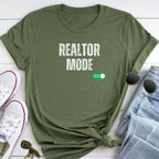 A green t-shirt with the text "REALTOR MODE" printed on it, along with a pair of sunglasses and a pair of sneakers, all laid out on a white surface.