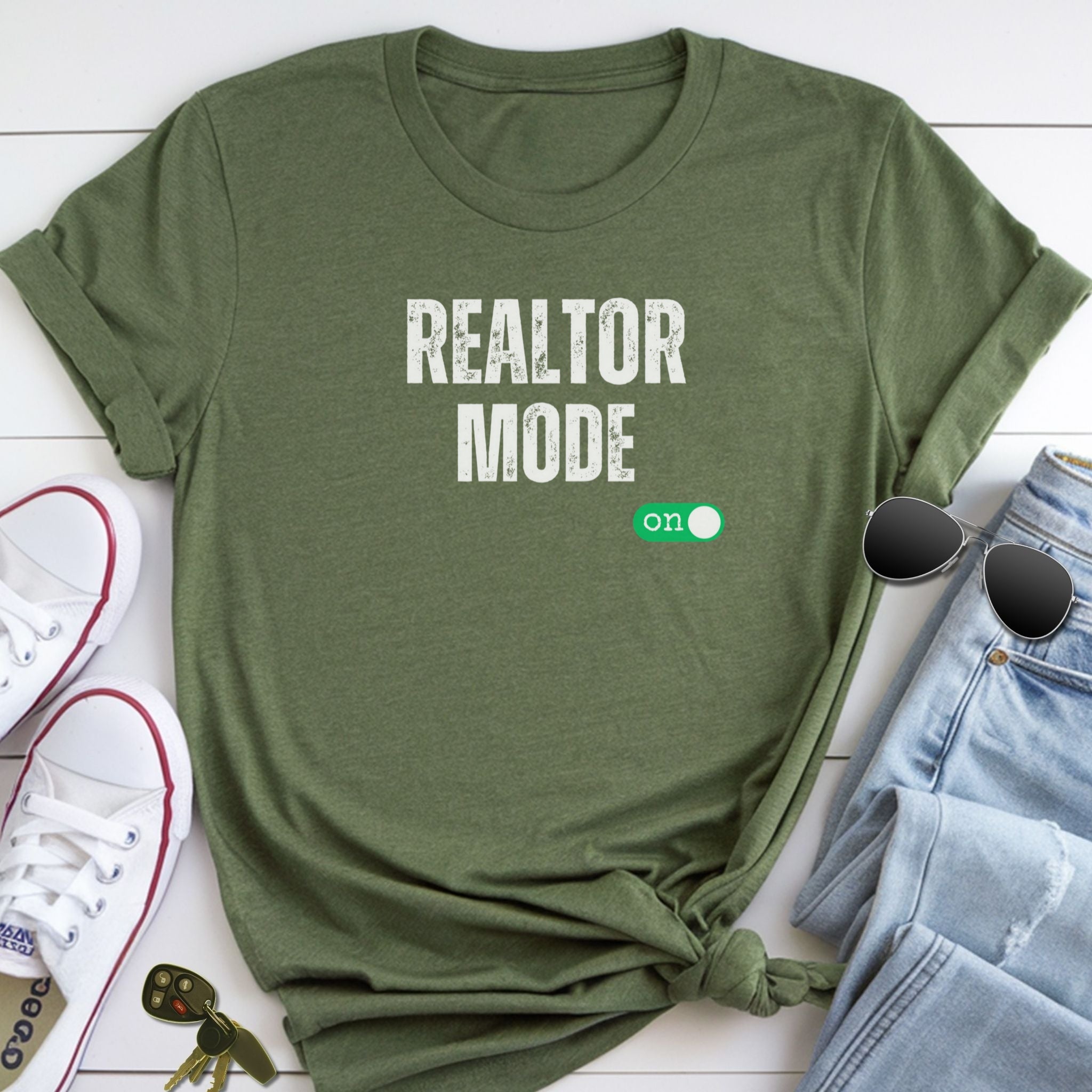 A green t-shirt with the text "REALTOR MODE" printed on it, along with a pair of sunglasses and a pair of sneakers, all laid out on a white surface.