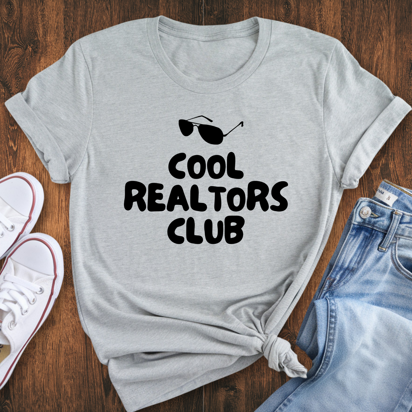 A gray t-shirt with the text "COOL REALTORS CLUB" printed on it, along with sunglasses and a casual outfit of jeans and sneakers.