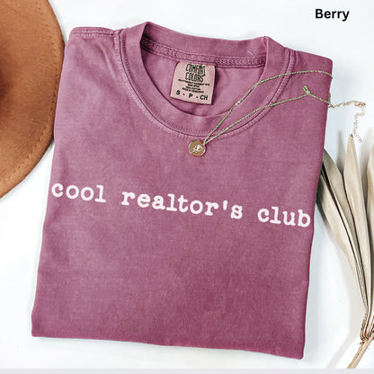 A pink t-shirt with the text "cool realtor's club" printed on it, along with a necklace and some leaves.