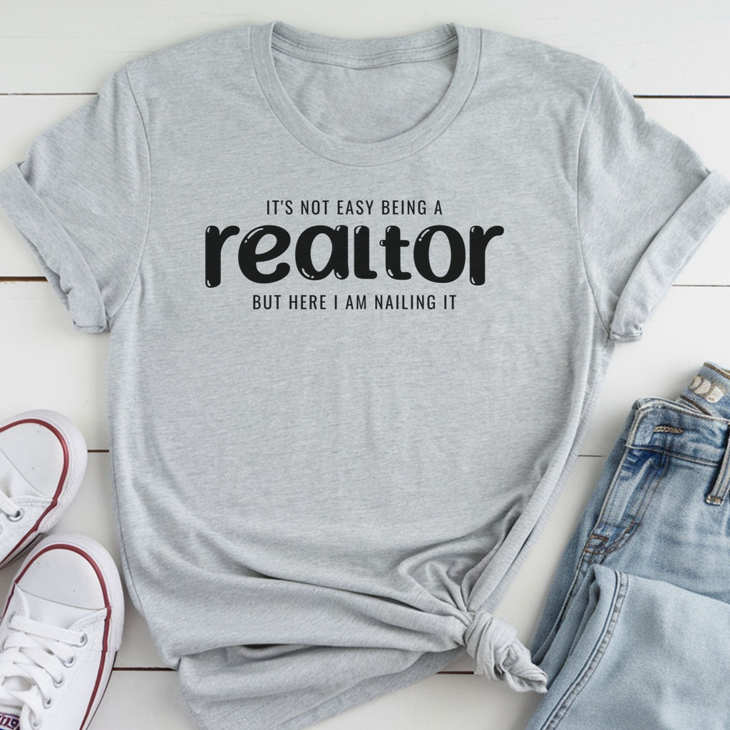 A gray t-shirt with the text "It's not easy being a realtor but here I am nailing it" printed on it, along with a pair of jeans and white sneakers.