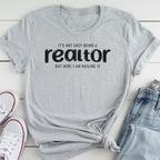 A gray t-shirt with the text "It's not easy being a realtor but here I am nailing it" printed on it, along with a pair of jeans and white sneakers.