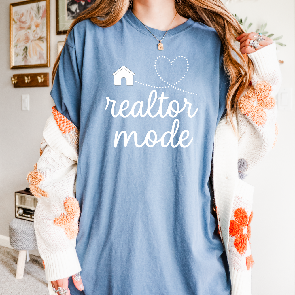 A person wearing a blue t-shirt with the text "realtor mode" printed on it, standing in a room with a floral patterned wall.