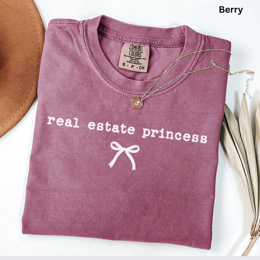 A pink t-shirt with the text "real estate princess" printed on it, along with a small white bow symbol. The t-shirt is folded and placed on a white surface, accompanied by a brown hat and some dried leaves.