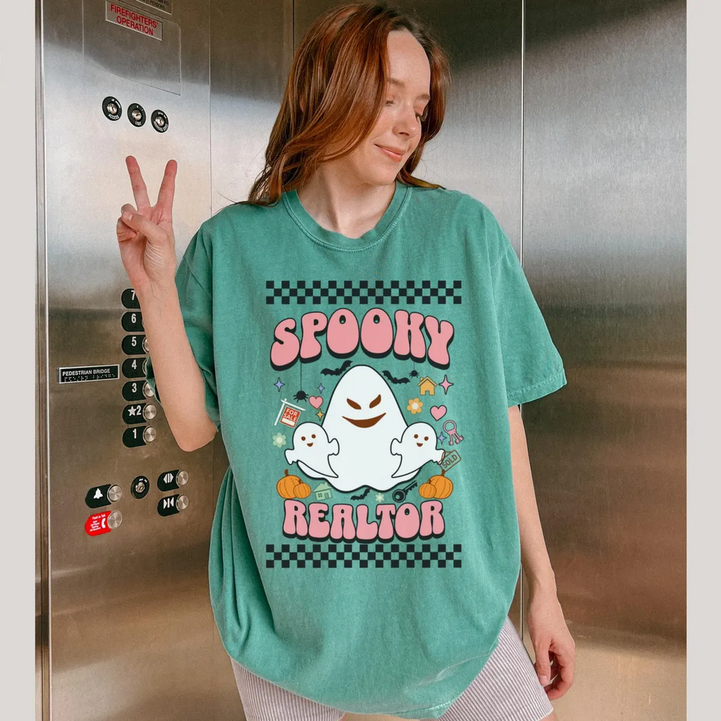 A woman wearing a green t-shirt with a graphic design of a ghost and the text "SPOKEY REALTOR" is posing in front of an elevator.