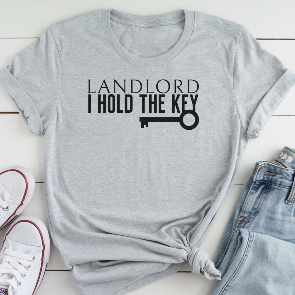 A gray t-shirt with the text "LANDLORD I HOLD THE KEY" printed on it, along with a key graphic. The t-shirt is placed on a white surface, and there are a pair of white sneakers and a pair of blue jeans visible in the image.