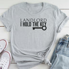 A gray t-shirt with the text "LANDLORD I HOLD THE KEY" printed on it, along with a key graphic. The t-shirt is placed on a white surface, and there are a pair of white sneakers and a pair of blue jeans visible in the image.