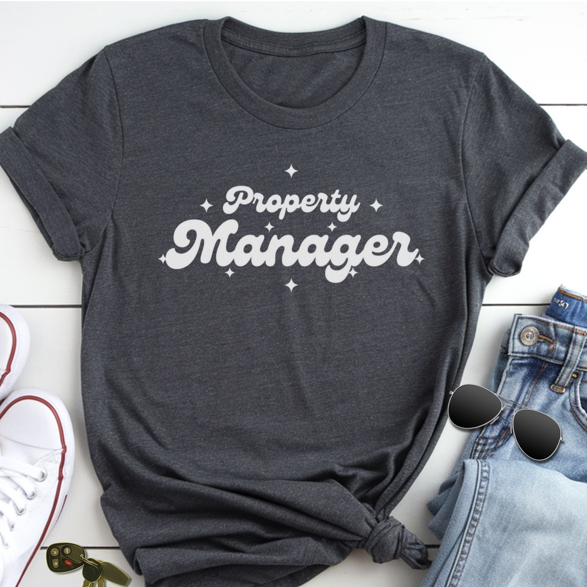 A gray t-shirt with the text "Property Manager" printed on it, along with some jeans and sunglasses, all laid out on a white surface.