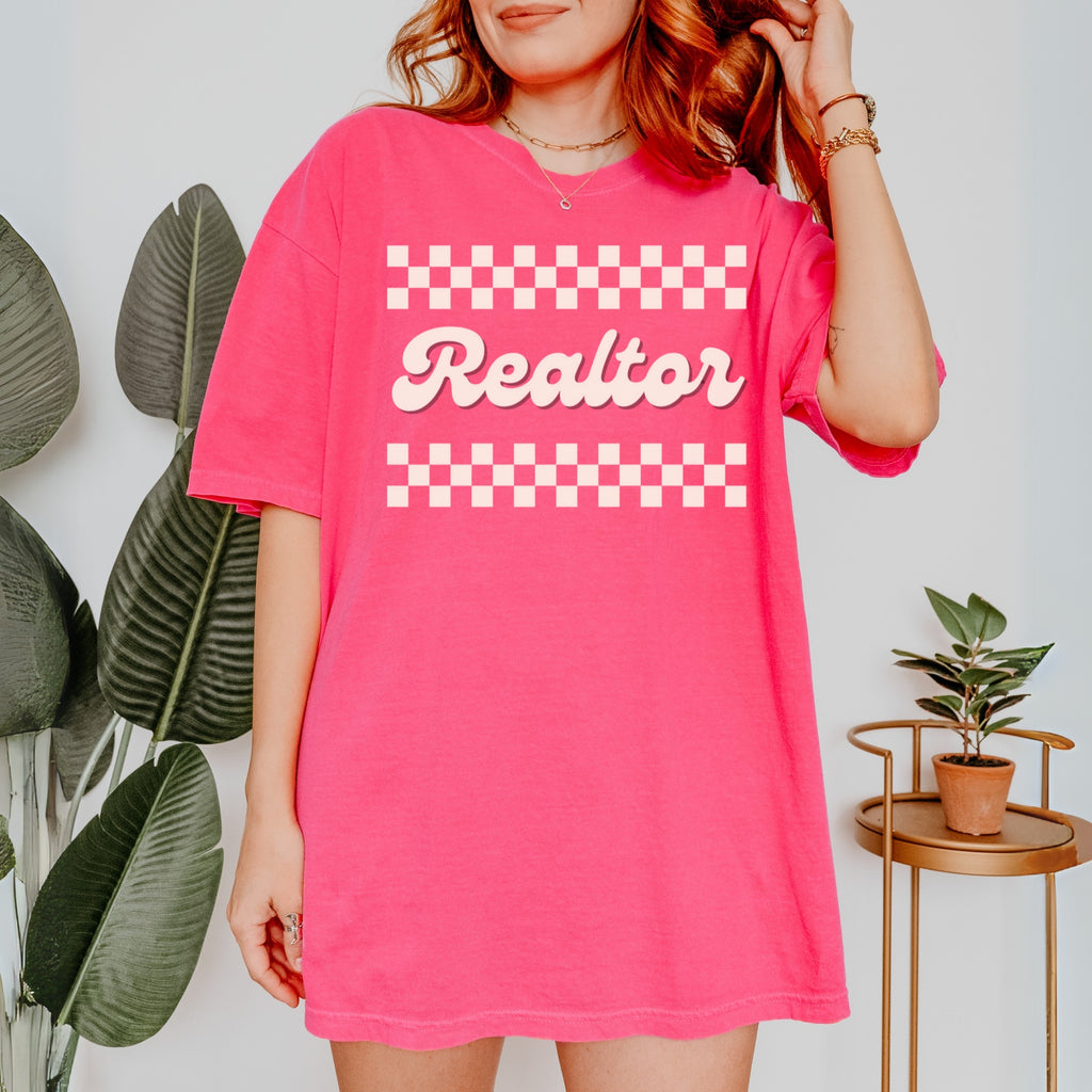 A woman wearing a pink t-shirt with the word "Realtor" printed on it, standing in front of a plant and a small table.