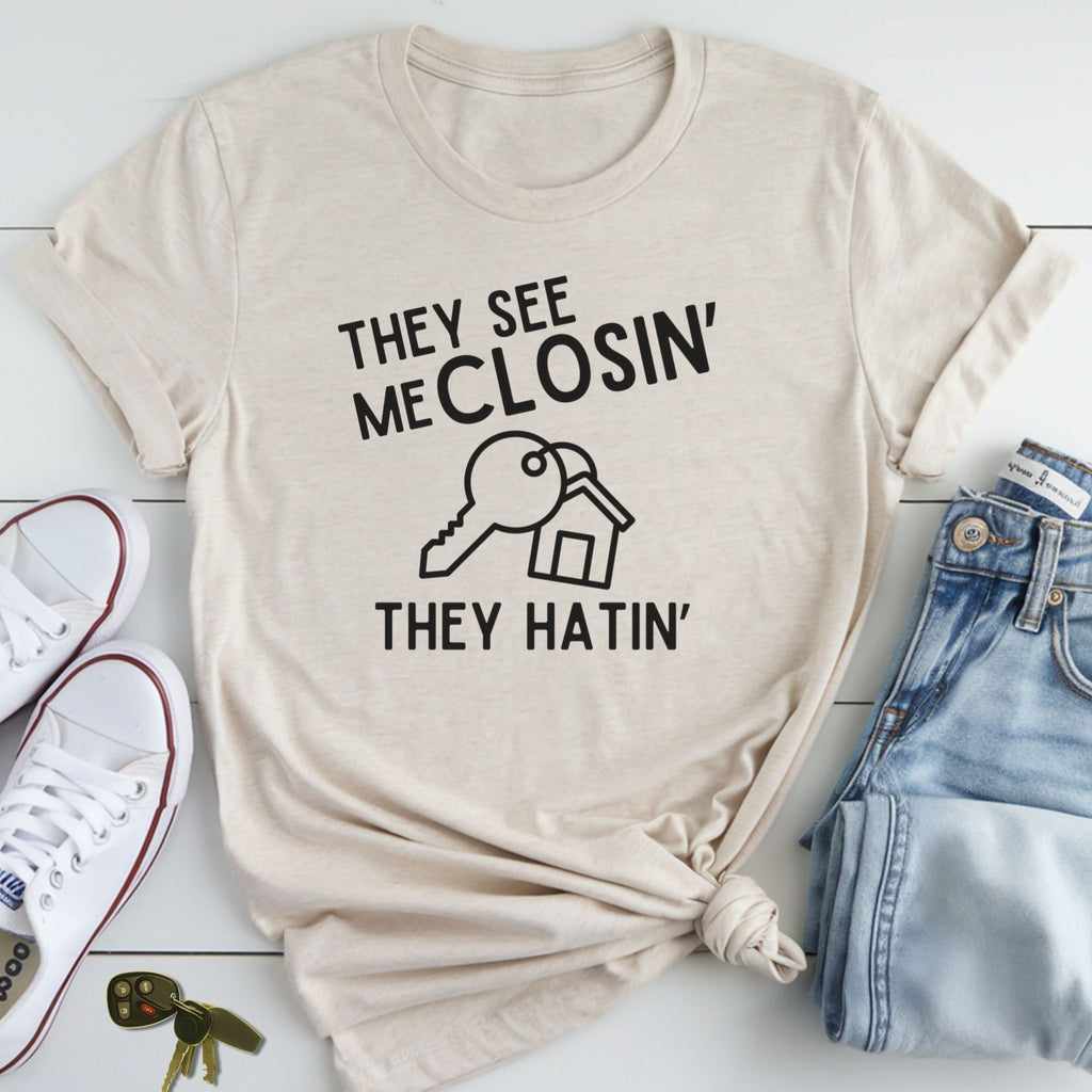 A beige t-shirt with a humorous text printed on it, along with a pair of jeans and sneakers, all laid out on a white surface.