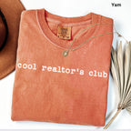 A red t-shirt with the text "cool realtor's club" printed on it, along with a gold necklace and a brown hat.