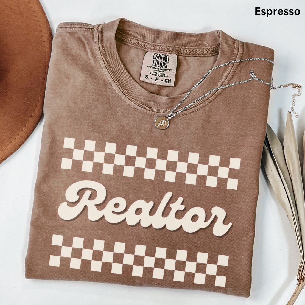 a brown t-shirt with a checkered pattern and the word "Realtor" printed on it. The t-shirt is laid out on a white surface, with a brown hat and some leaves visible in the background.