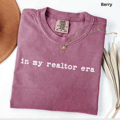 A pink t-shirt with the text "in my realtor era" printed on it, along with a necklace and a hat.