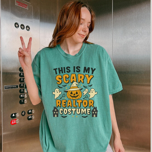 A woman wearing a green t-shirt with a Halloween-themed design, including a jack-o-lantern, bats, and ghostly figures, is making a peace sign with her right hand.