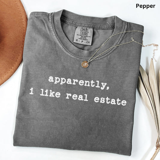 A gray t-shirt with the text "apparently, I like real estate" printed on it, along with a small pendant necklace.
