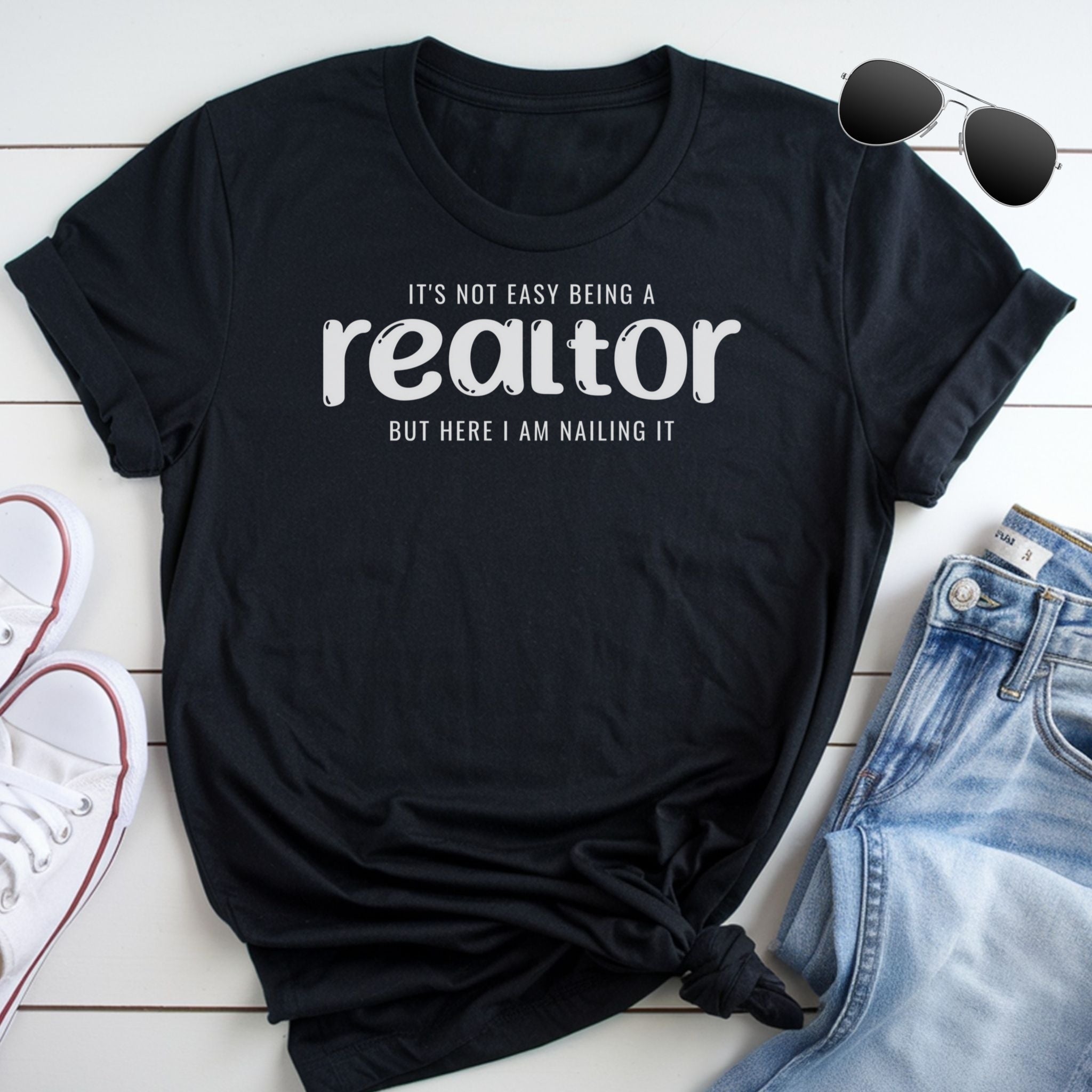 A black t-shirt with white text that reads "It's not easy being a realtor but here I am nailing it" is displayed on a white surface, accompanied by a pair of sunglasses and a pair of jeans.