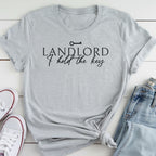 A gray t-shirt with the text "LANDLORD I hold the key" printed on it, along with a key symbol. The t-shirt is laid out on a white surface, and there are a pair of jeans and white sneakers visible in the image.