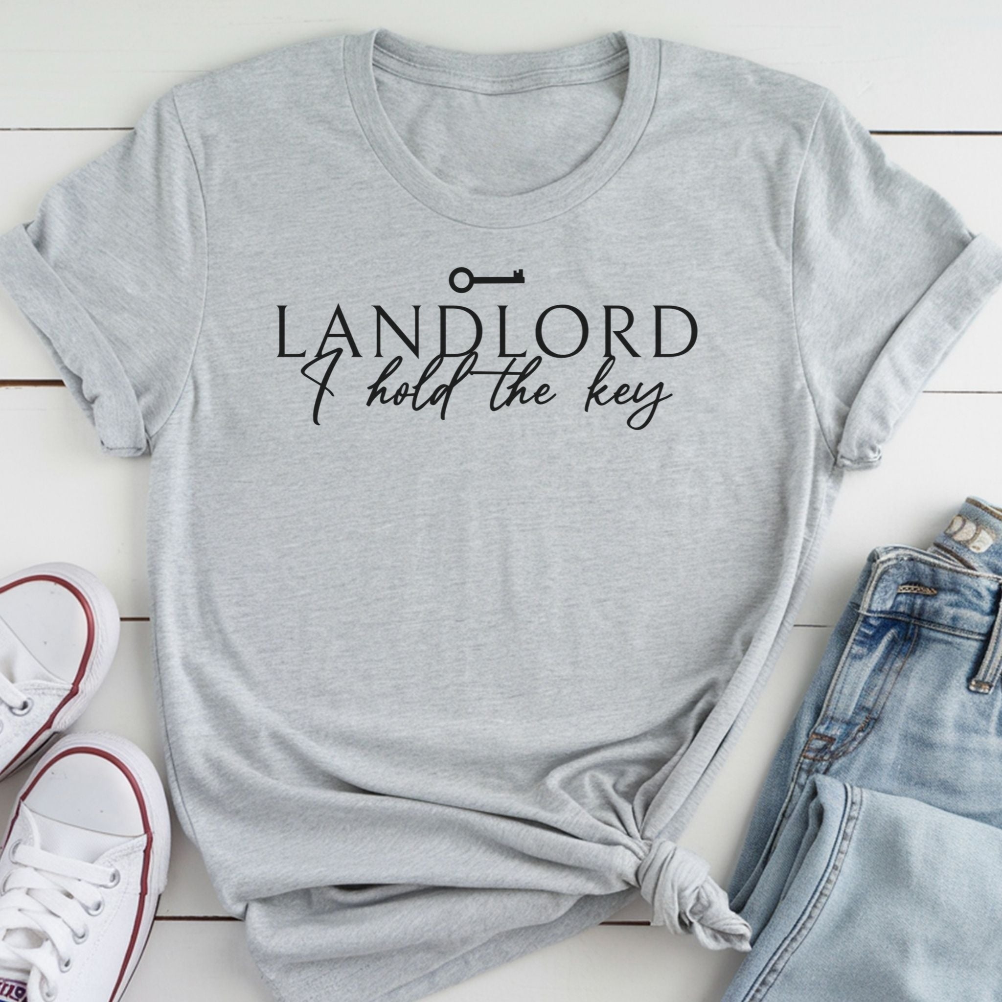 A gray t-shirt with the text "LANDLORD I hold the key" printed on it, along with a key symbol. The t-shirt is laid out on a white surface, and there are a pair of jeans and white sneakers visible in the image.