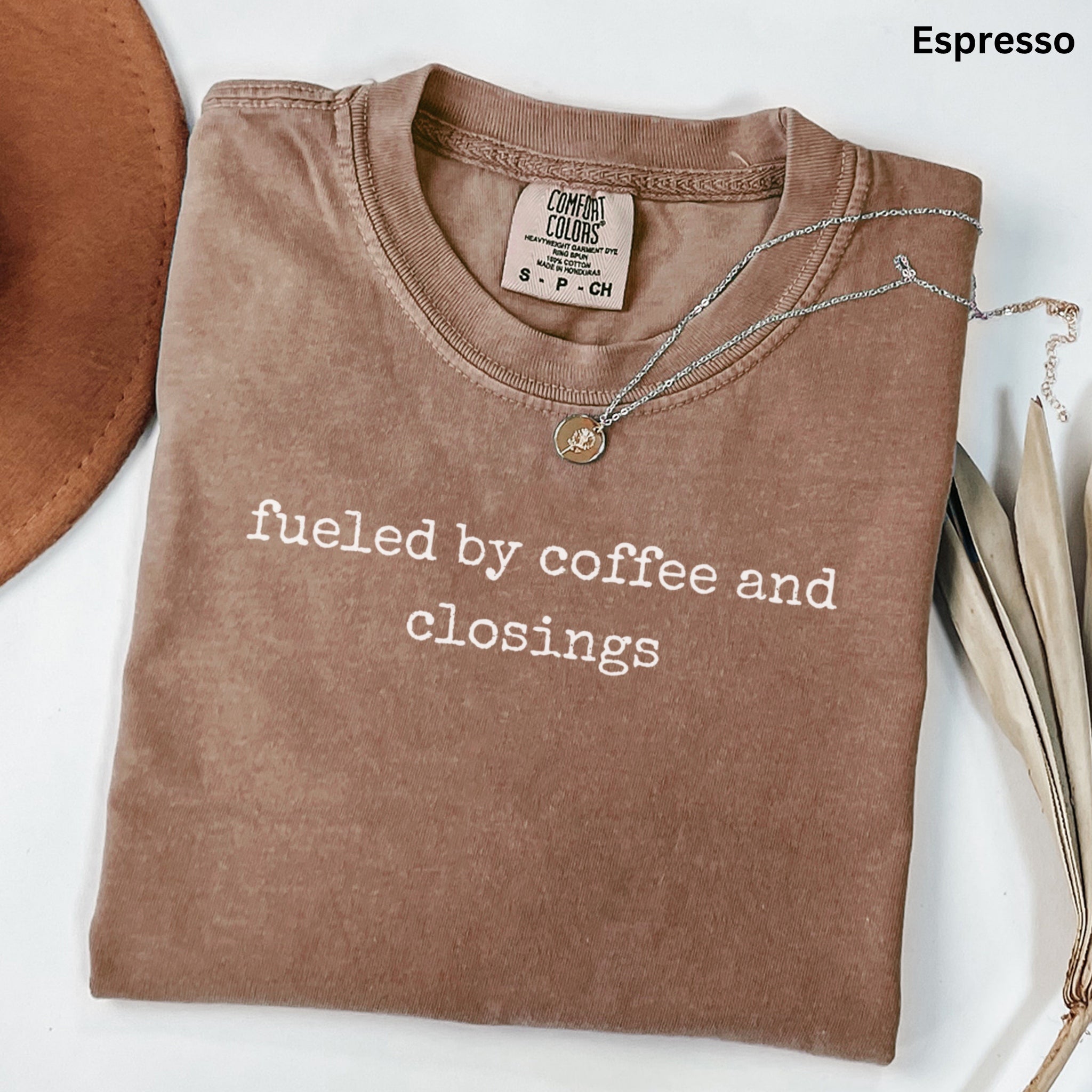 A brown t-shirt with the text "fueled by coffee and closings" printed on it, along with a necklace and some leaves.