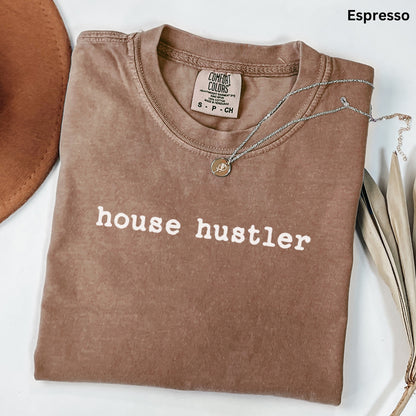 a brown t-shirt with the text "house hustler" printed on it, along with a necklace and some leaves.