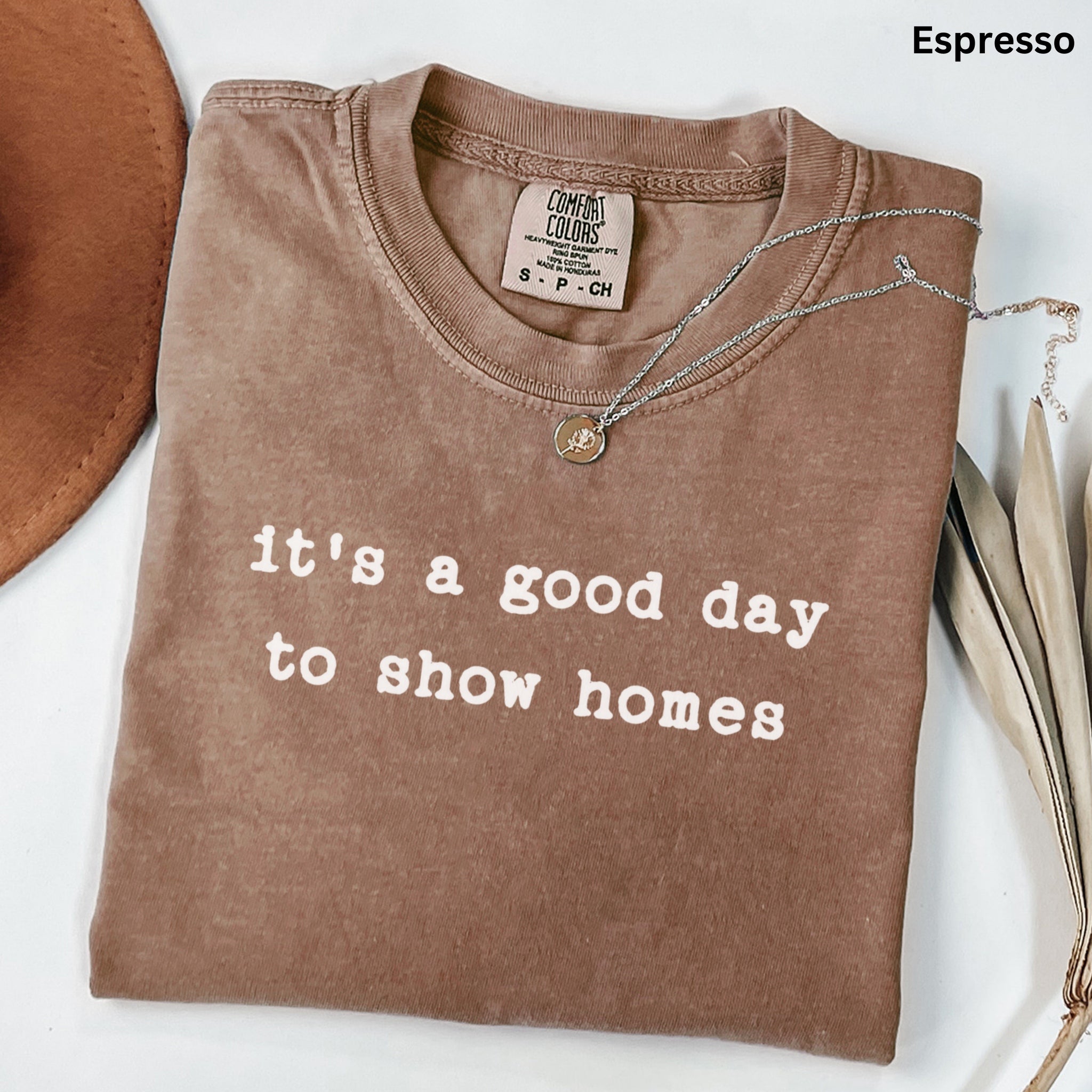 A brown t-shirt with the text "it's a good day to show homes" printed on it, along with a necklace and some leaves.