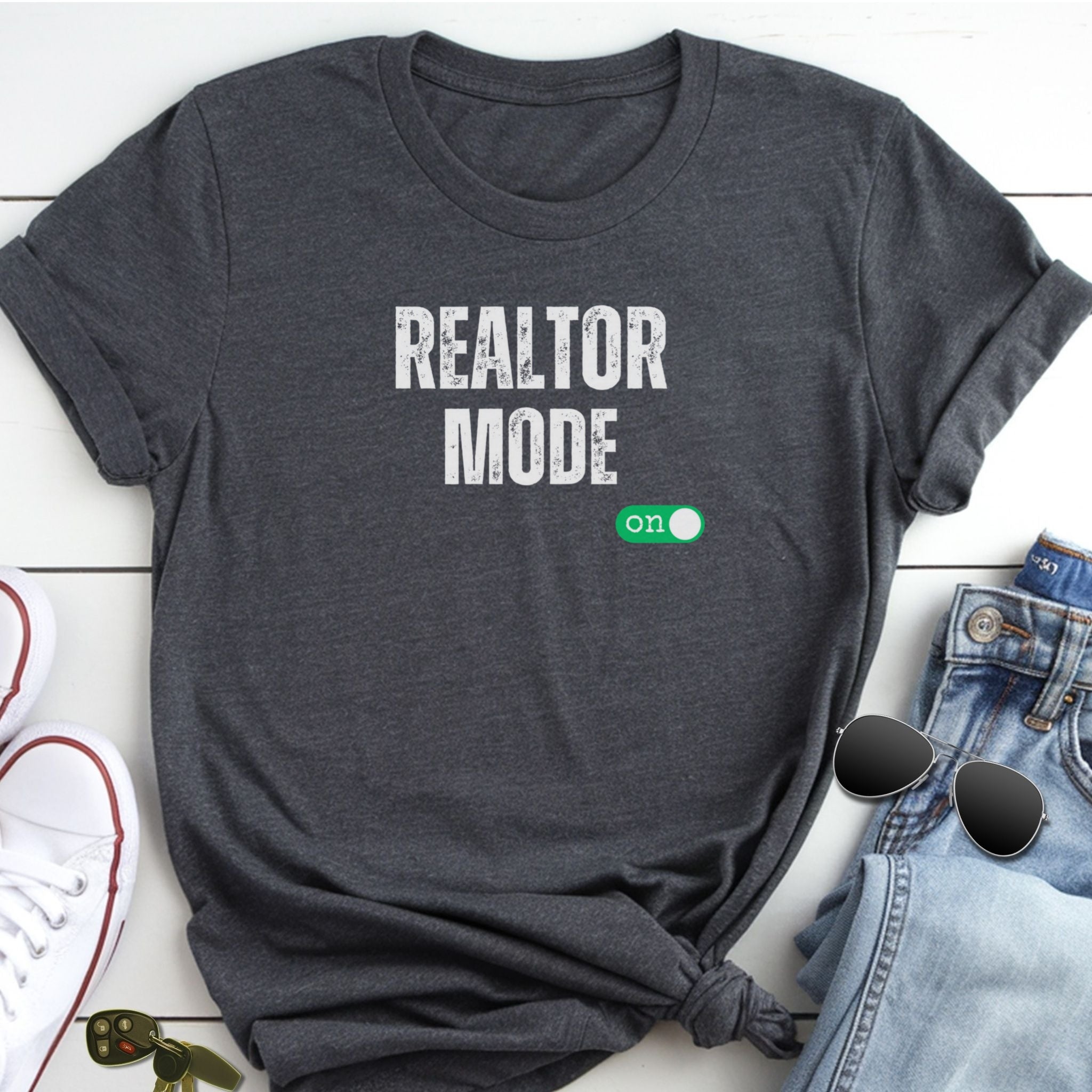 A gray t-shirt with the text "REALTOR MODE" printed on it, along with a green button that says "ON". The t-shirt is placed on a white surface, and there are some jeans and sunglasses visible in the background.