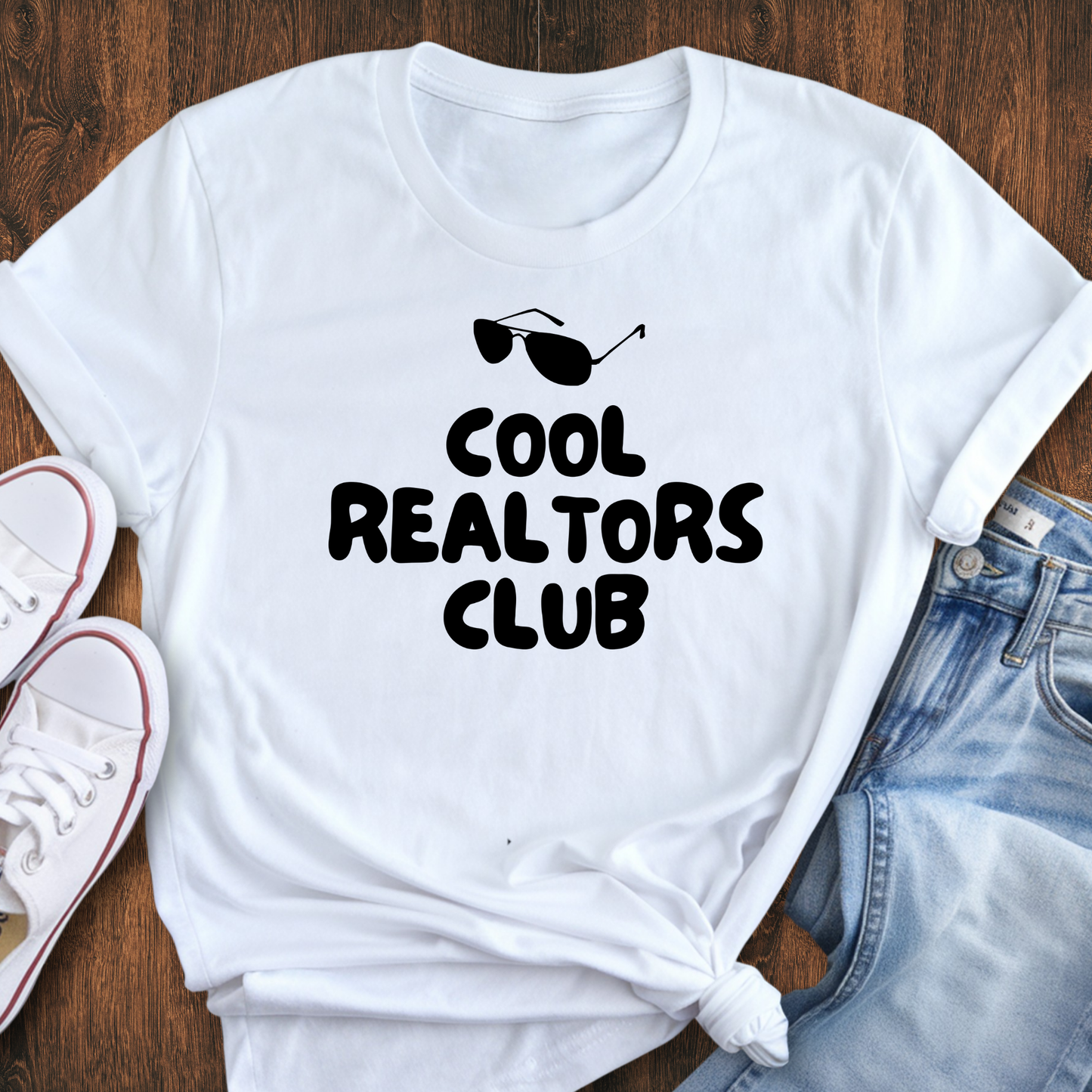 A white t-shirt with the text "COOL REALTORS CLUB" printed on it, along with sunglasses and a casual outfit consisting of jeans and white sneakers.
