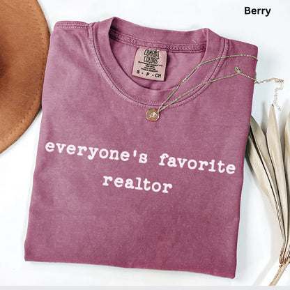 A pink t-shirt with the text "Everyone's favorite realtor" printed on it, along with a small gold pendant. The t-shirt is folded and placed on a white surface, with a brown hat and some leaves visible in the background.