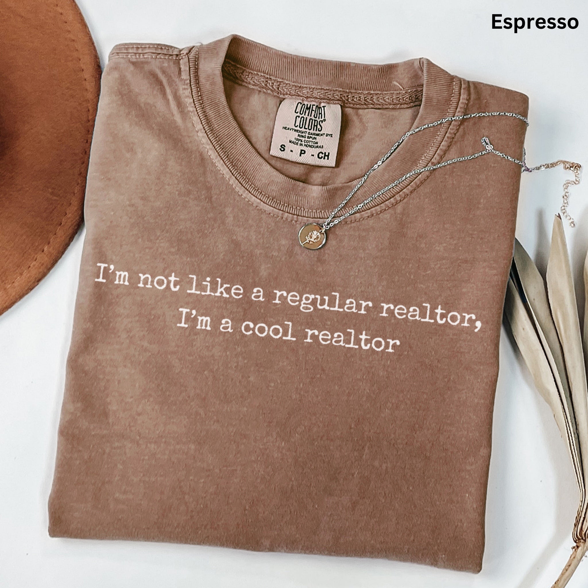 a brown t-shirt with a humorous text printed on it, along with a necklace and some leaves.
