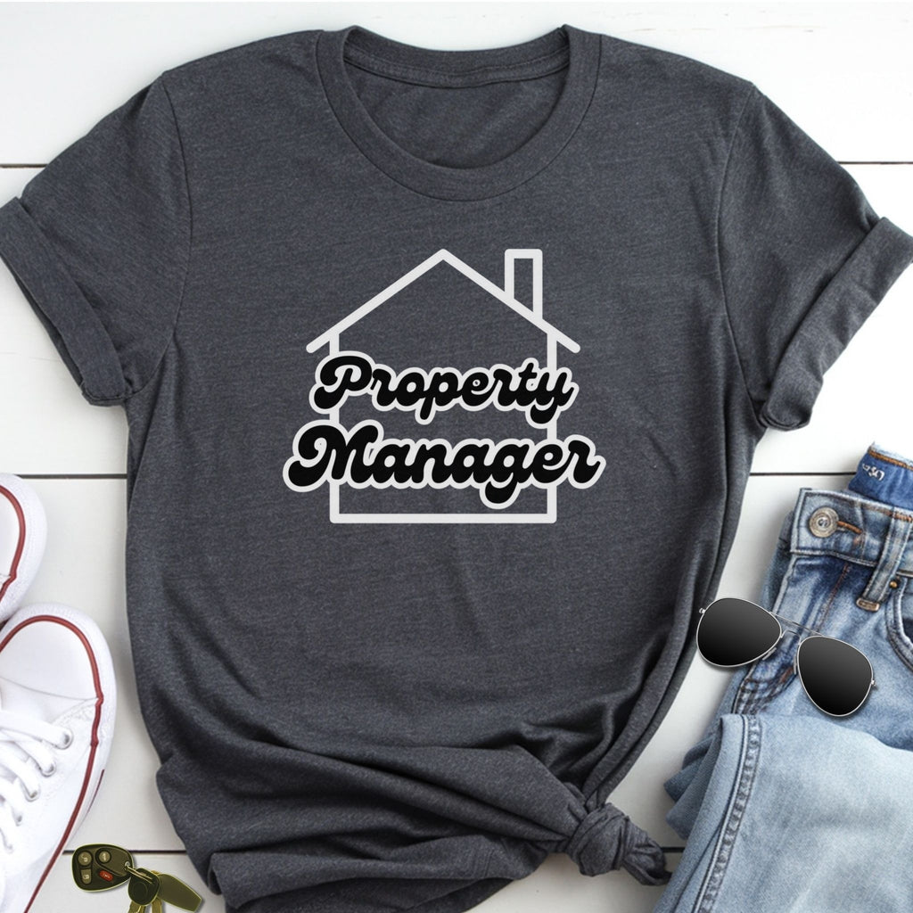 A gray t-shirt with the text "Property Manager" printed on it, along with a graphic of a house. The t-shirt is placed on a white surface, and there are some jeans and sunglasses visible in the background.