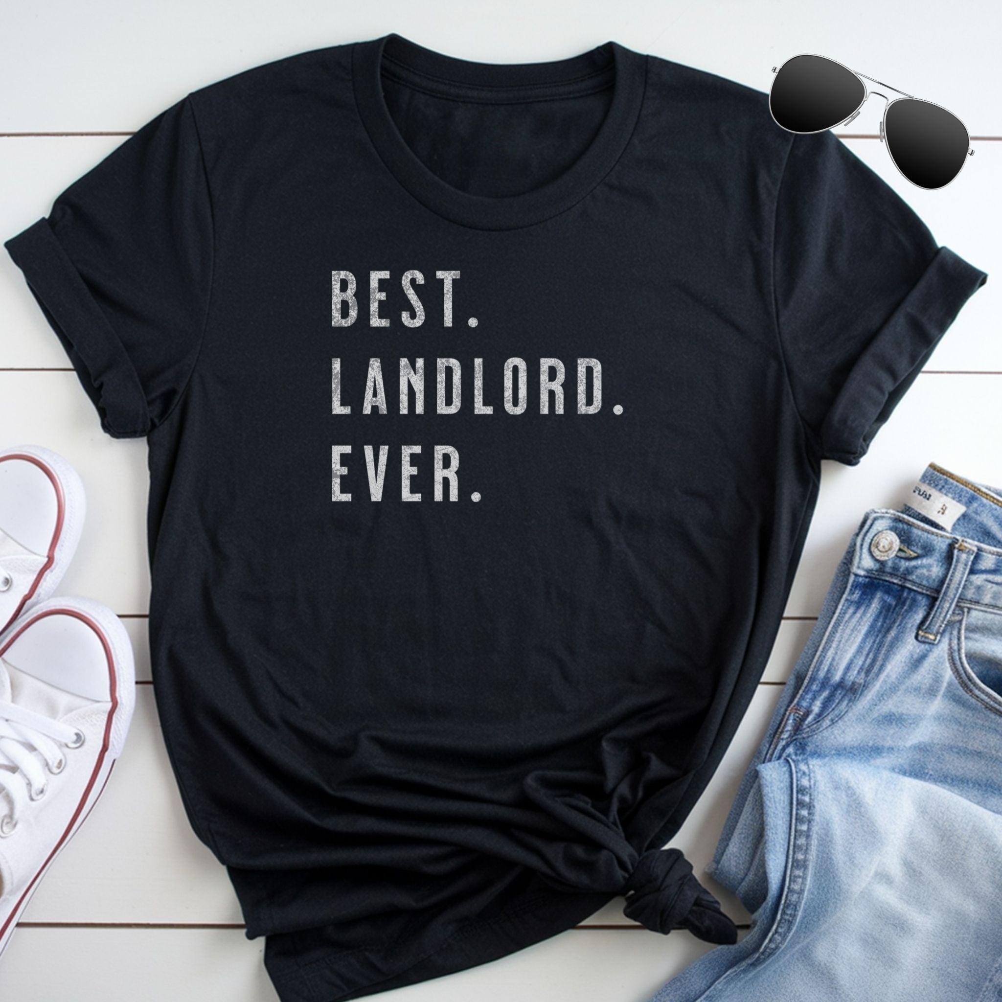 Black t-shirt with 'Best Landlord Ever' text on a wooden surface with jeans and sneakers.