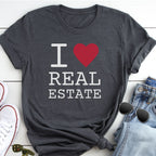 A gray t-shirt with the text "I ♥ REAL ESTATE" printed on it, along with a red heart symbol. The t-shirt is placed on a white surface, and there are some sunglasses and a pair of jeans visible in the background.