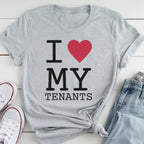A gray t-shirt with the text "I ♥ MY TENANTS" printed on it, along with a red heart symbol. The t-shirt is placed on a white surface, and there are a pair of jeans and white sneakers visible in the background.