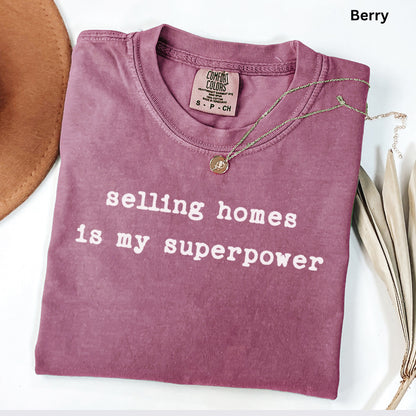 A purple t-shirt with the text "selling homes is my superpower" printed on it, along with a necklace and a hat.