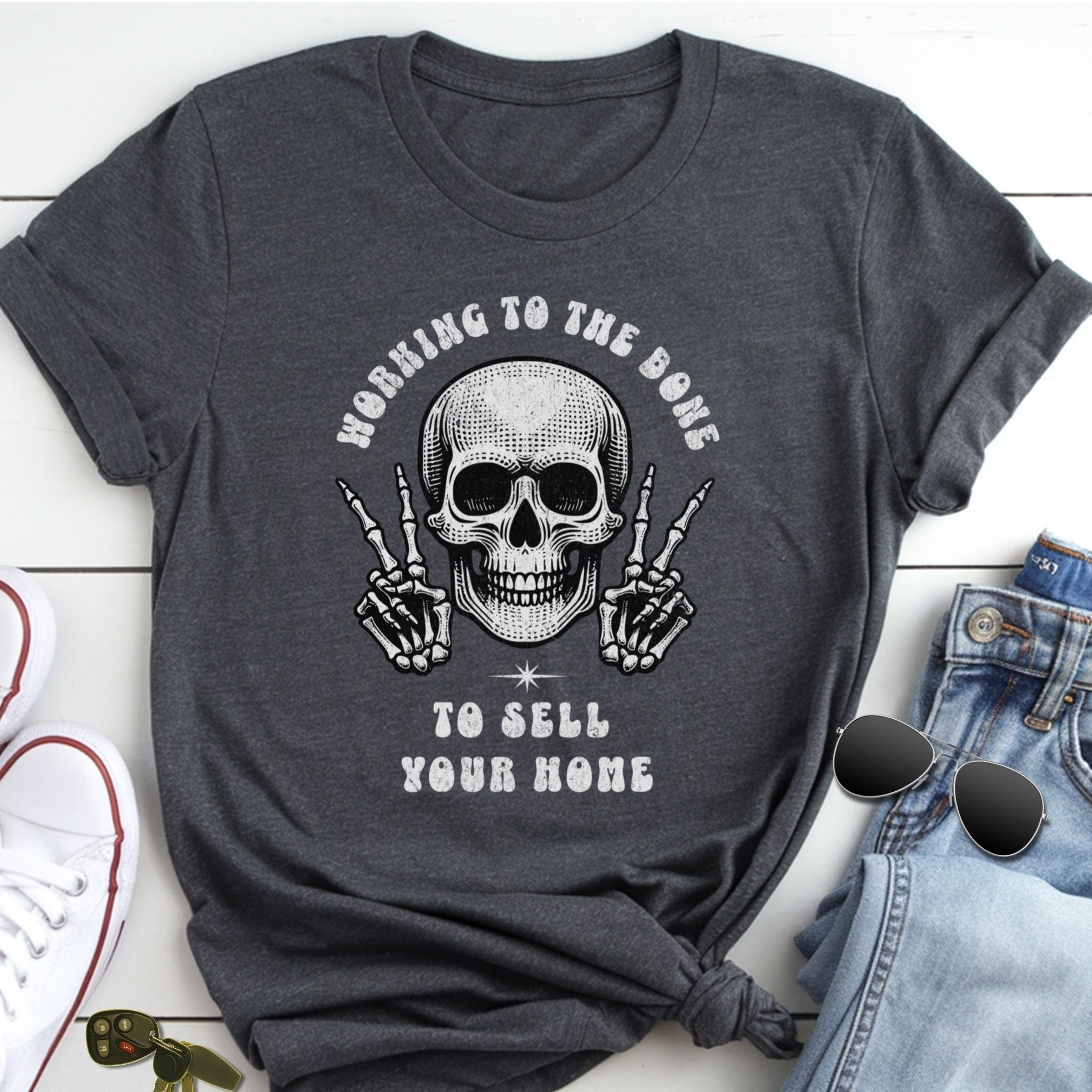 A gray t-shirt with a skull graphic and text, along with a pair of jeans and sunglasses, placed on a white surface.
