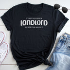 A black t-shirt with white text that reads "IT'S NOT EASY BEING A LANDLORD BUT HERE I AM NAILING IT" is laid out on a white surface, accompanied by a pair of sunglasses and a pair of jeans.
