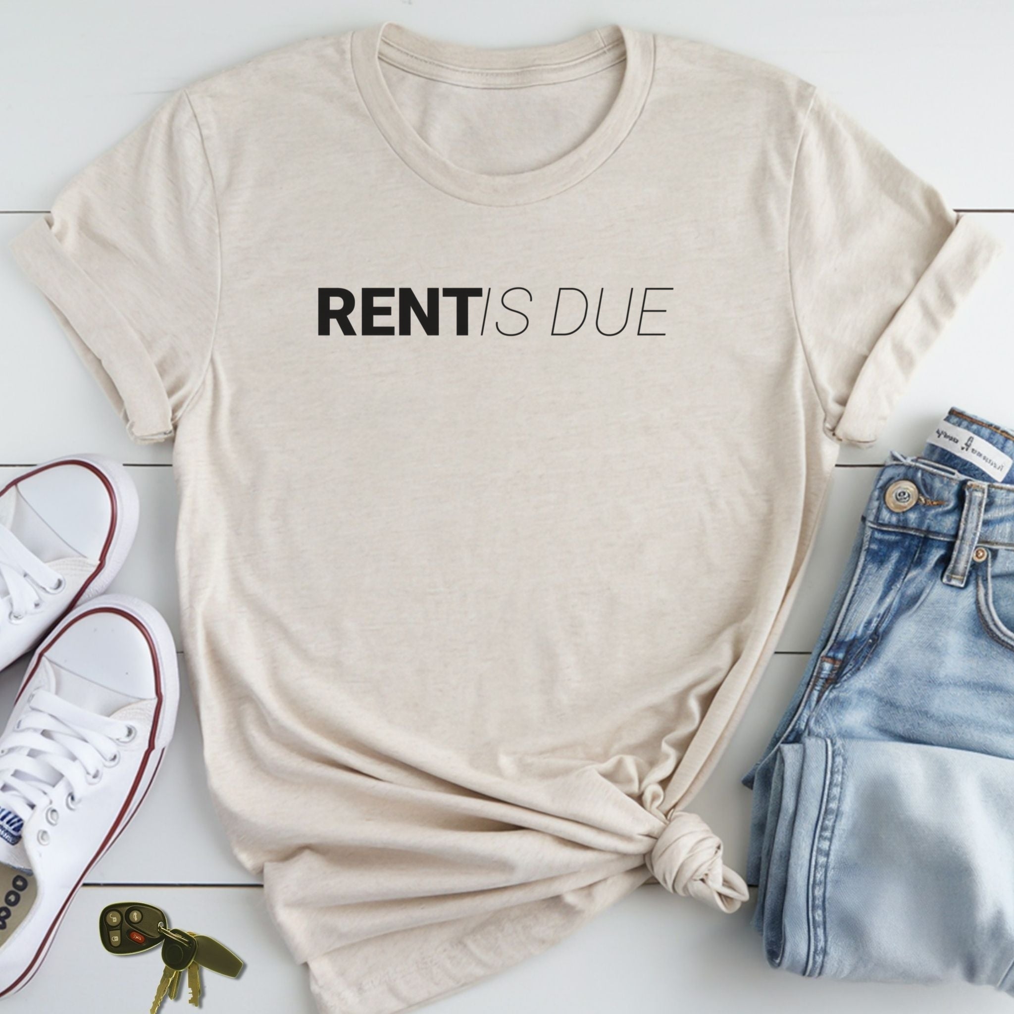 A beige t-shirt with the text "RENT IS DUE" printed on it, along with a pair of jeans and white sneakers, all laid out on a white surface.