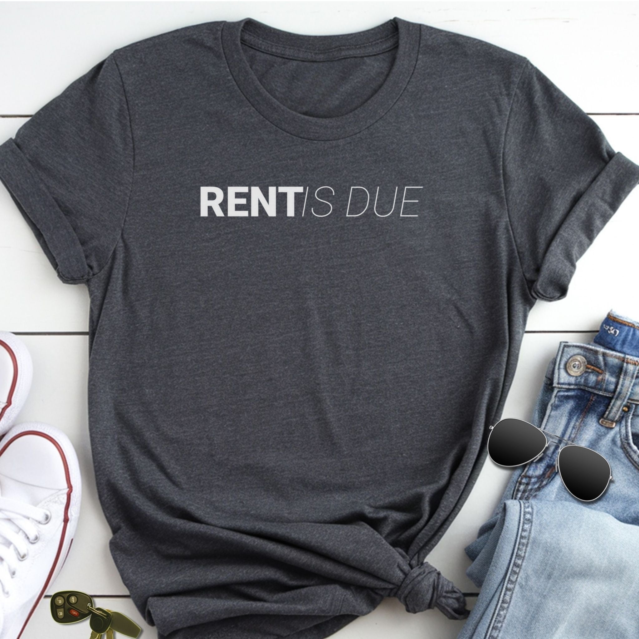 A gray t-shirt with the text "RENT IS DUE" printed on it, along with a pair of jeans and sunglasses, all laid out on a white surface.