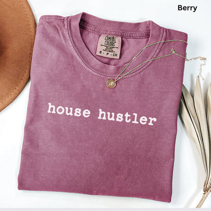 A pink t-shirt with the text "house hustler" printed on it, along with a necklace and a hat.
